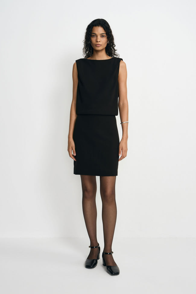 A model wearing a black sleeveless mini dress with a boat neck and draped bodice above a straight skirt, paired with black ankle-strap shoes.