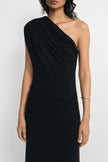 Close-up of a model in a black one-shoulder dress, showing folds gathered at the covered shoulder and sweeping diagonally across the bodice and waist.