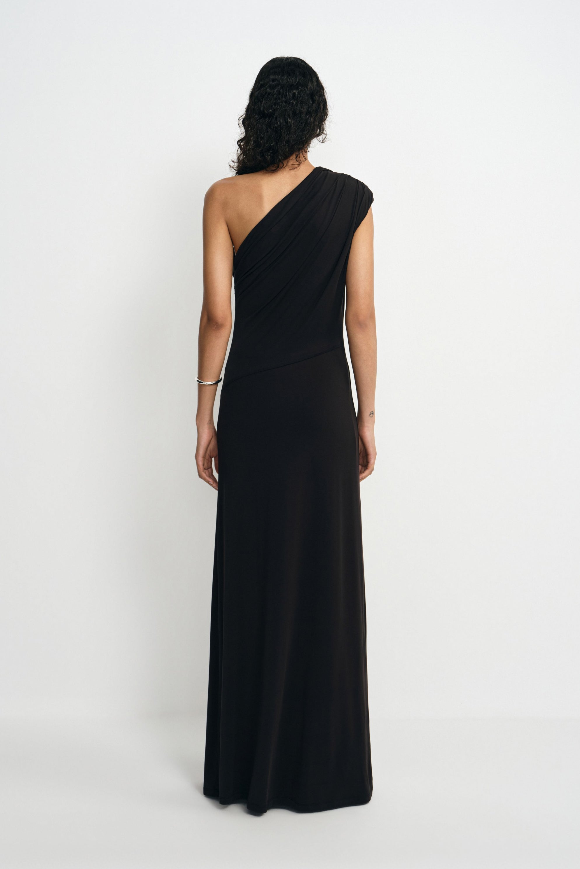 Back view of a model wearing a black maxi dress with a one-shoulder neckline and diagonal folds across the back above the long skirt.