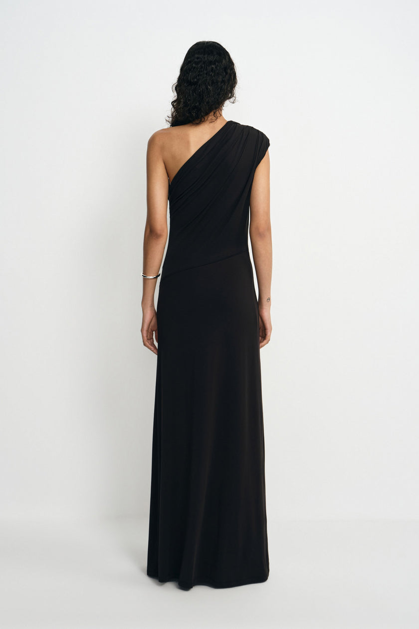 Back view of a model wearing a black maxi dress with a one-shoulder neckline and diagonal folds across the back above the long skirt.