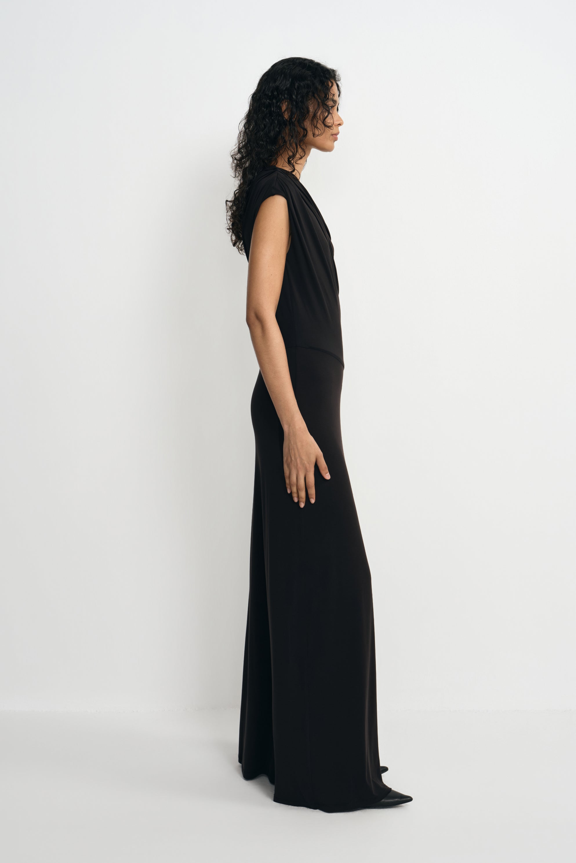 Side view of a model in a black one-shoulder maxi dress, showing draping from the covered shoulder and a long skirt falling close to the floor.