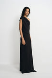 Three-quarter view of a model wearing a black maxi dress with a draped one-shoulder bodice and long column skirt, with pointed black shoes beneath the hem.