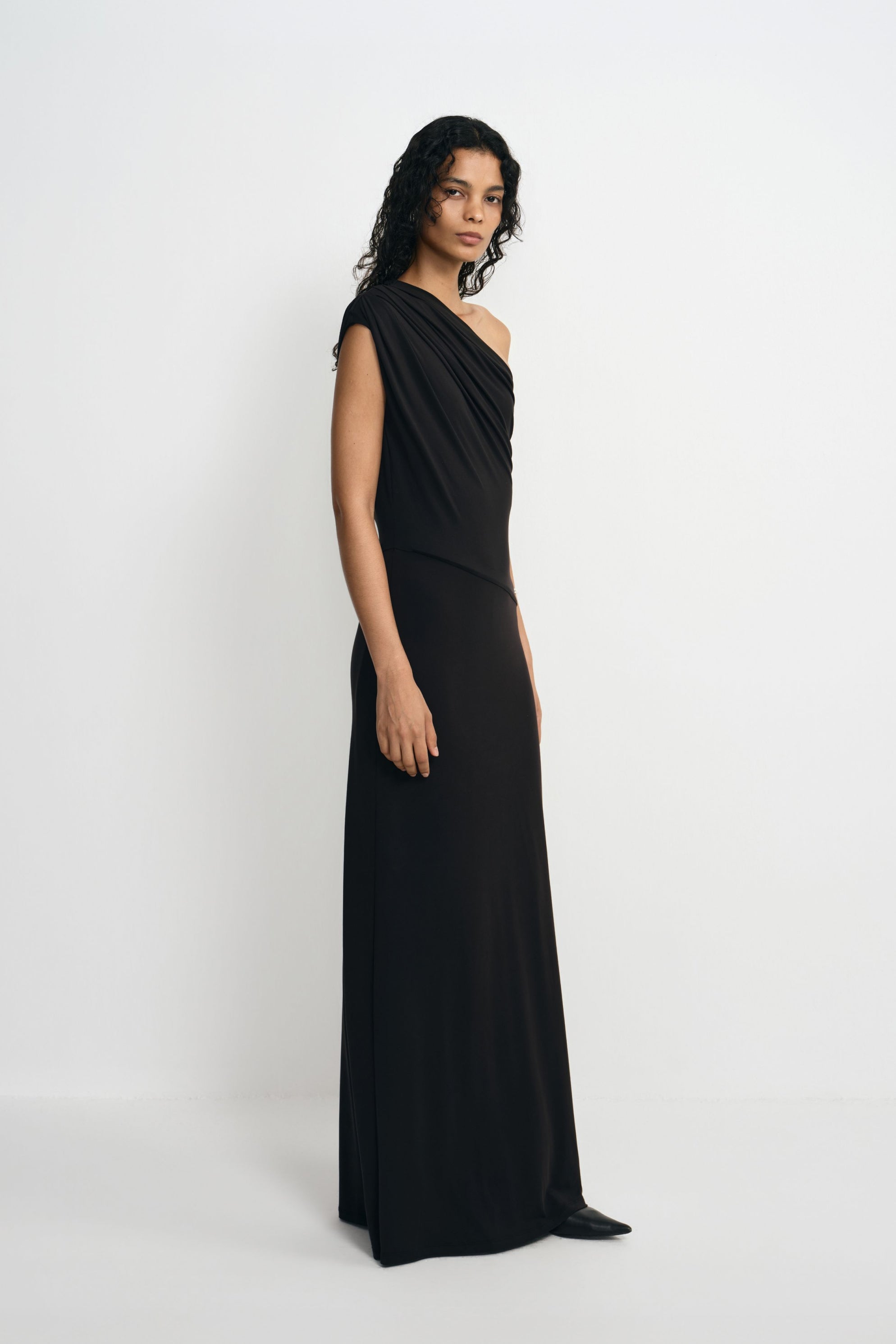 Three-quarter view of a model wearing a black maxi dress with a draped one-shoulder bodice and long column skirt, with pointed black shoes beneath the hem.