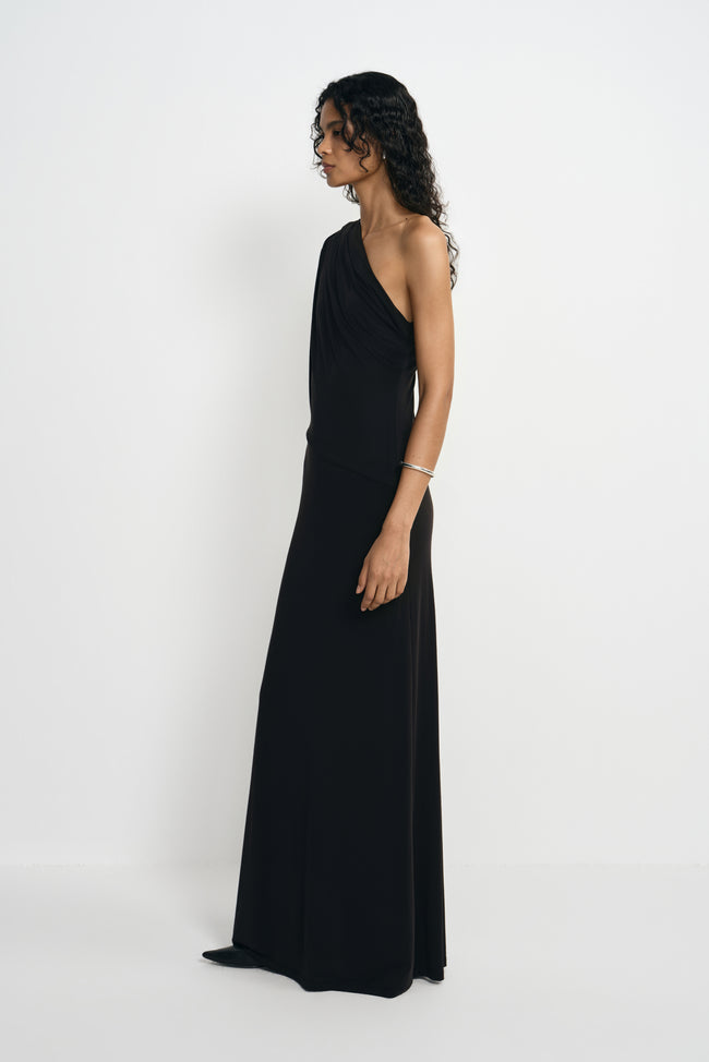 Side view of a model in a black maxi dress from the bare shoulder, showing the diagonal neckline and waist draping, styled with a silver cuff.