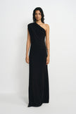 A model wearing a black one-shoulder maxi dress with diagonal draping across the bodice and waist, paired with pointed black shoes and a silver cuff.