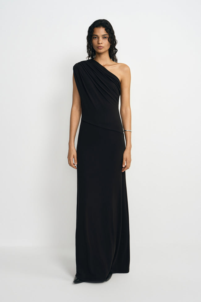 A model wearing a black one-shoulder maxi dress with diagonal draping across the bodice and waist, paired with pointed black shoes and a silver cuff.
