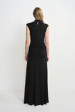 Back view of a model in a black maxi dress, showing a small keyhole below the high neckline, gathered waist and hips, and a long flared skirt.