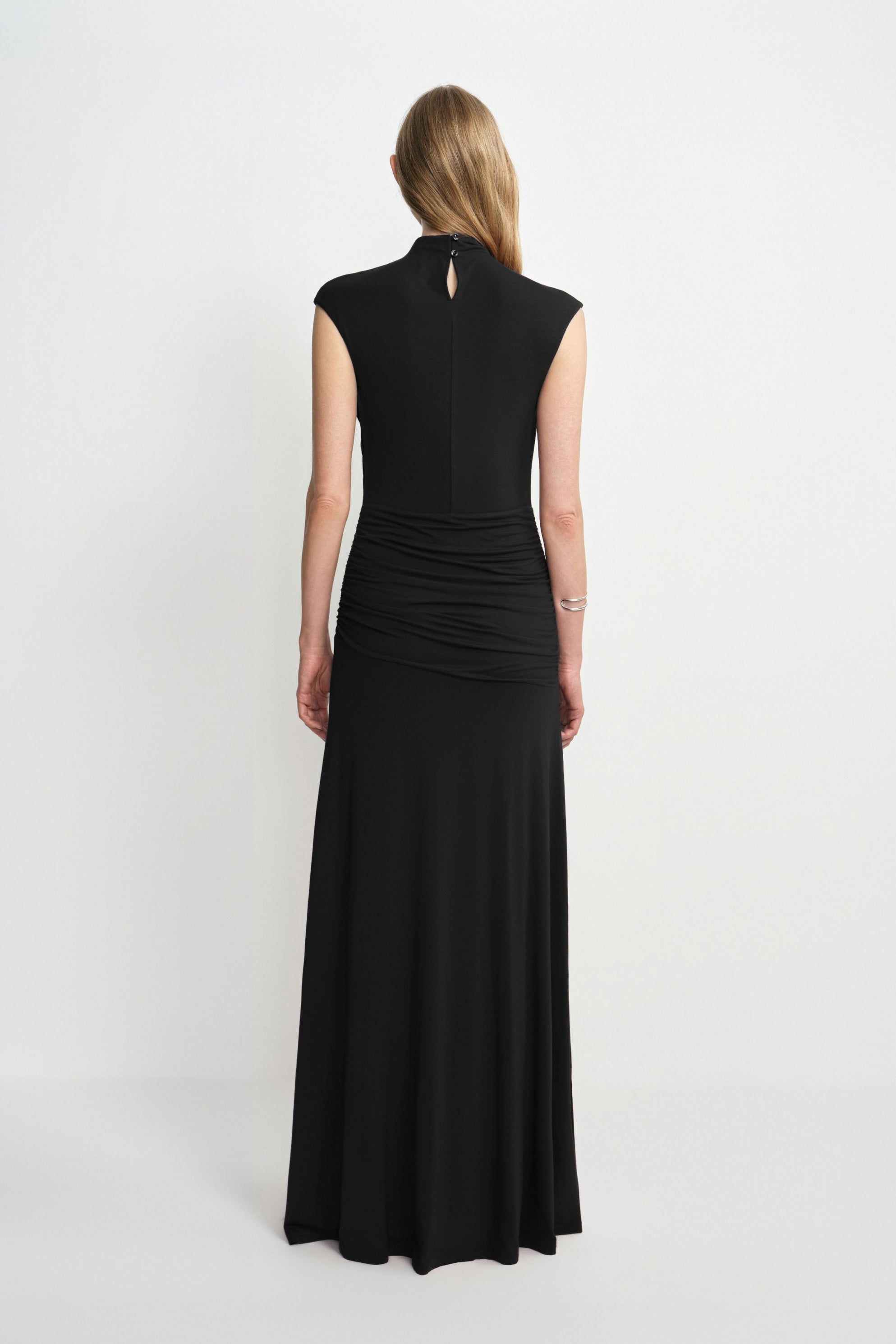 Back view of a model in a black maxi dress, showing a small keyhole below the high neckline, gathered waist and hips, and a long flared skirt.