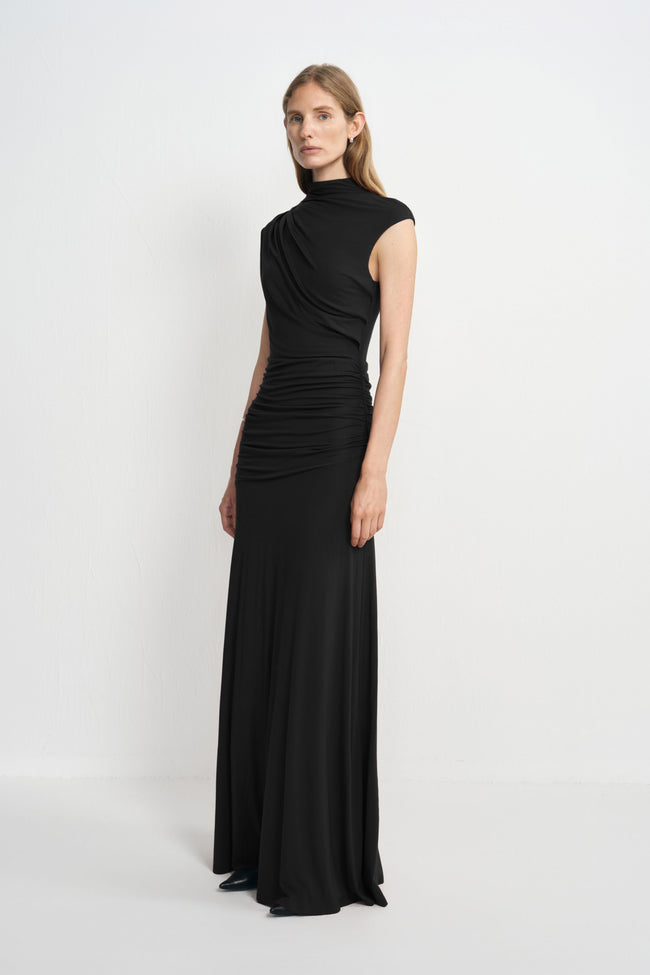 Three-quarter view of a model wearing a black maxi dress with a high neckline and asymmetric bodice draping, paired with pointed black shoes.