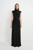 A model facing forward in a black maxi dress with a high neck, diagonal bodice folds and ruched hips, styled with a silver cuff.