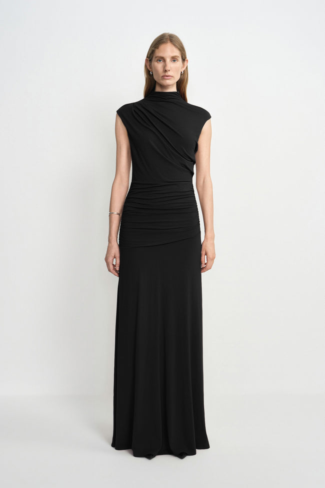 A model facing forward in a black maxi dress with a high neck, diagonal bodice folds and ruched hips, styled with a silver cuff.