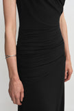 Close-up of a black dress on a model, showing diagonal folds gathering at the side of the waist and continuing across the hips beside a silver cuff.