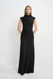 A model wearing a black high-neck maxi dress with diagonal folds across the bodice and gathers through the waist and hips, with black shoes beneath the hem.
