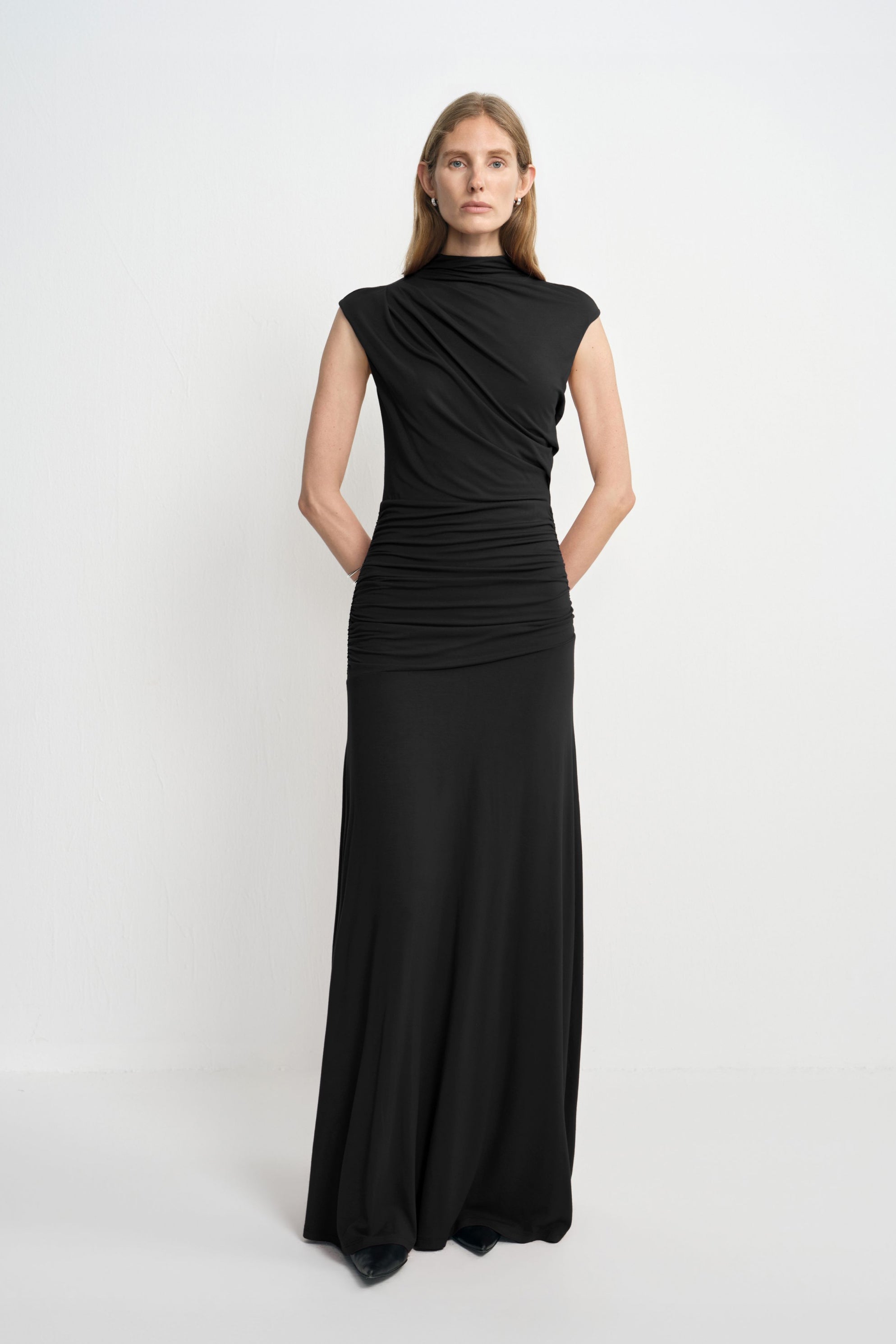 A model wearing a black high-neck maxi dress with diagonal folds across the bodice and gathers through the waist and hips, with black shoes beneath the hem.