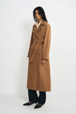 A woman modeling a long camel-brown belted trench coat over a pale yellow collared shirt and dark wide-leg trousers, standing against a plain light gray studio background.