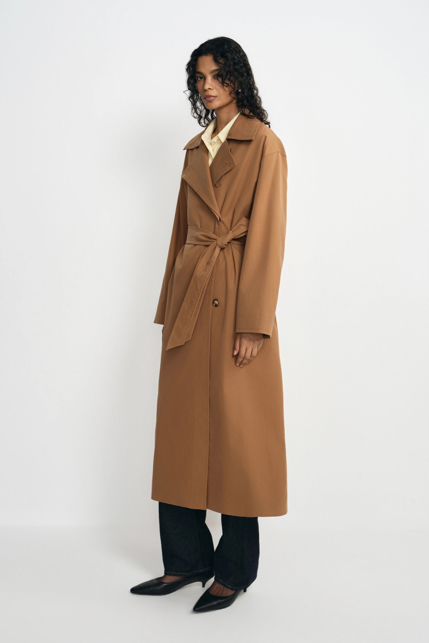 A woman modeling a long camel-brown belted trench coat over a pale yellow collared shirt and dark wide-leg trousers, standing against a plain light gray studio background.