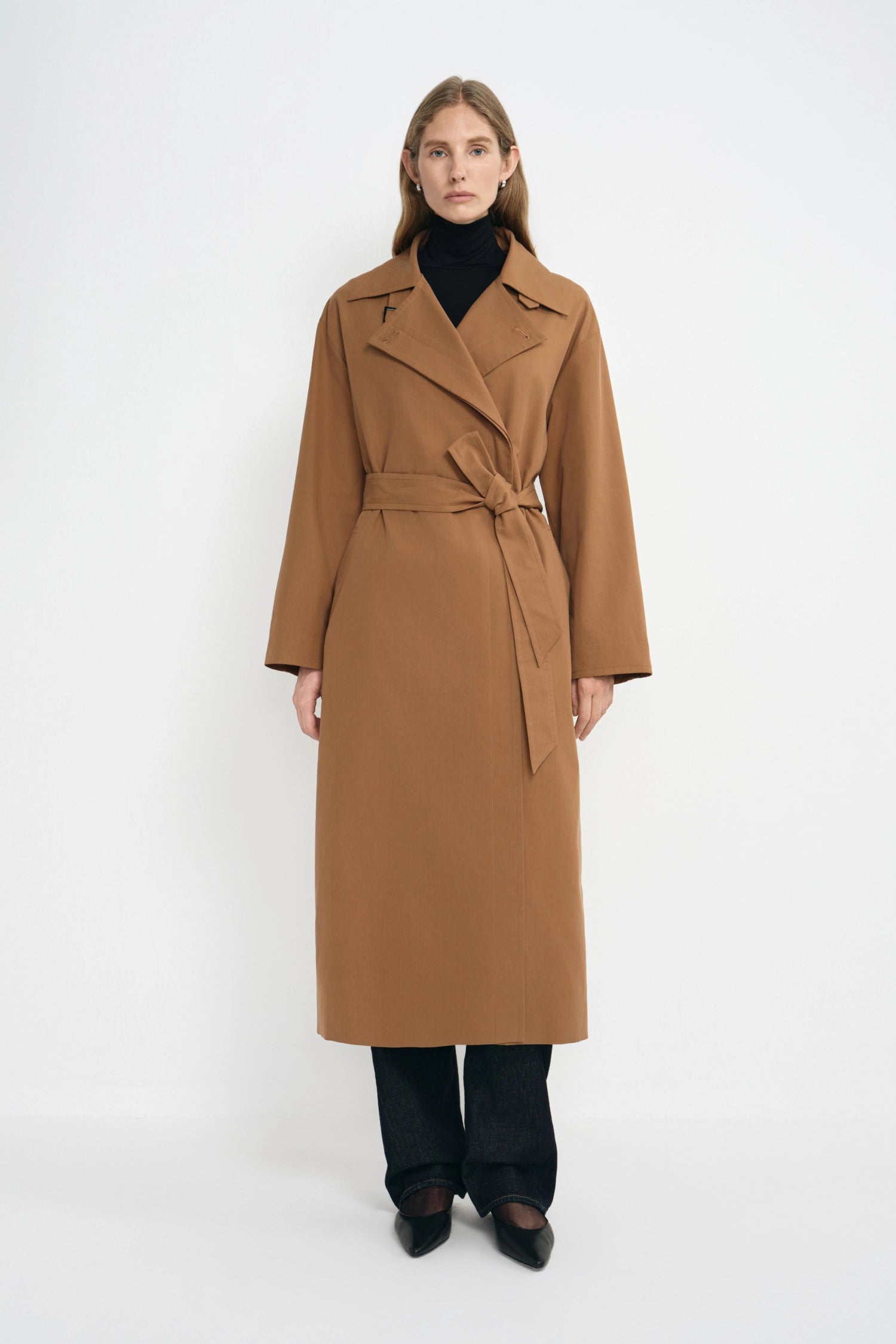A woman stands against a plain light gray studio background wearing a long camel-brown belted coat over a black turtleneck, black wide-leg pants, and black pointed shoes.