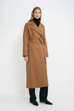 A woman modeling a long camel-brown trench coat with a tie waist belt, worn over a black turtleneck, black trousers, and black pointed shoes against a plain white studio background.