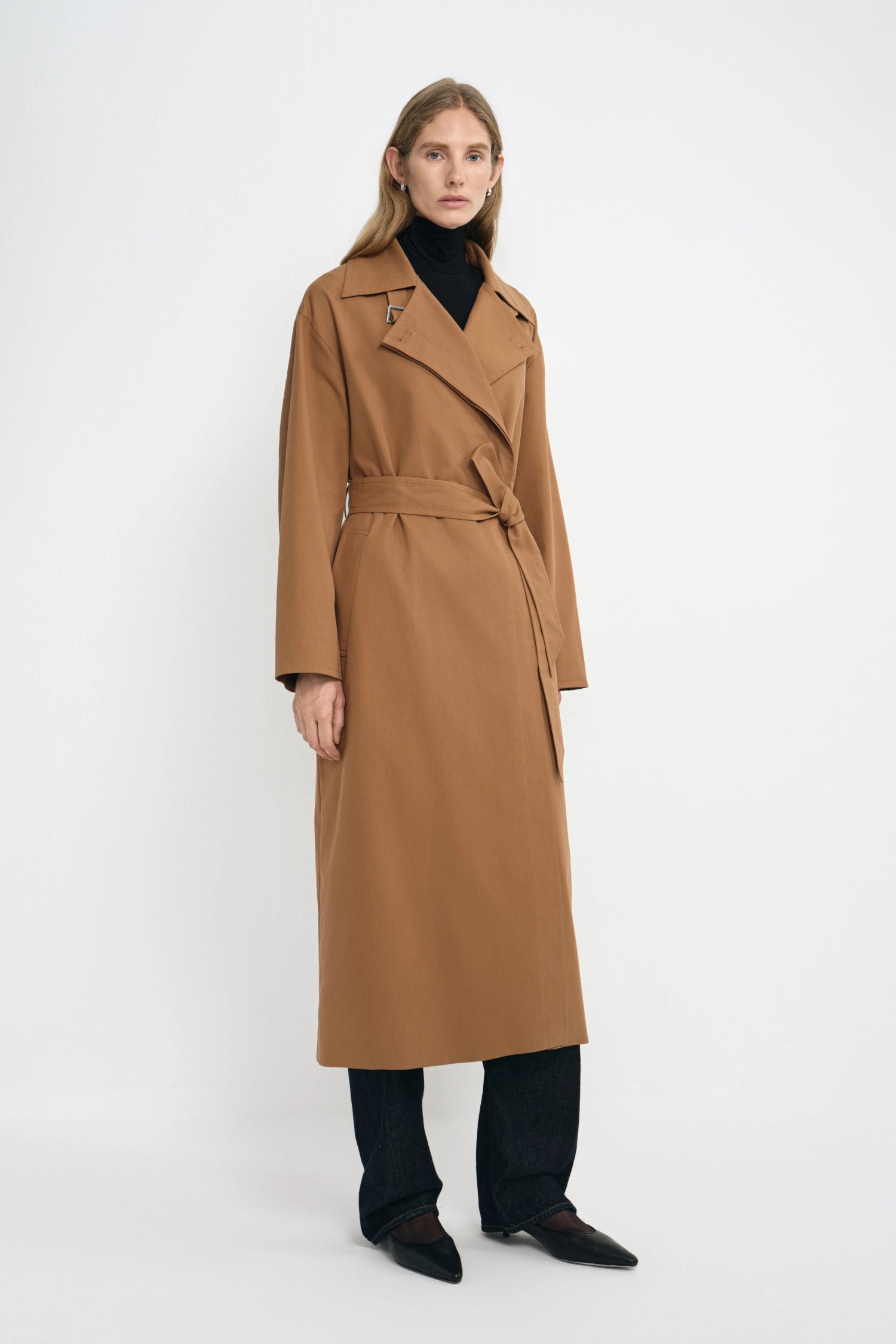 A woman modeling a long camel-brown trench coat with a tie waist belt, worn over a black turtleneck, black trousers, and black pointed shoes against a plain white studio background.