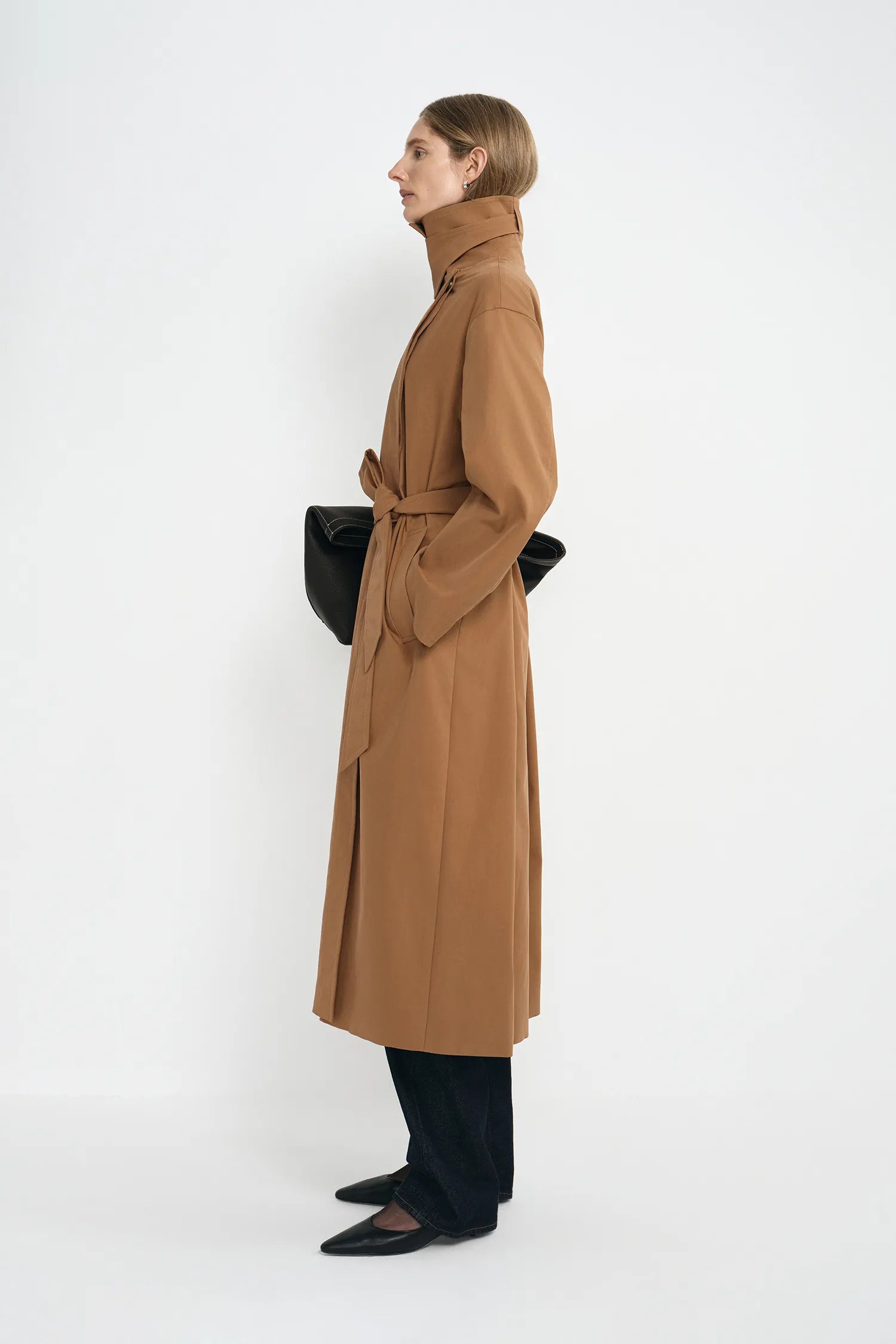 A model wearing a coppery brown oversized trench coat with a self-tie belt, side view.