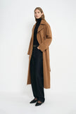 A model wearing a coppery brown oversized trench coat, pairing a black turtleneck and dark wash jeans, turned slightly to the side.