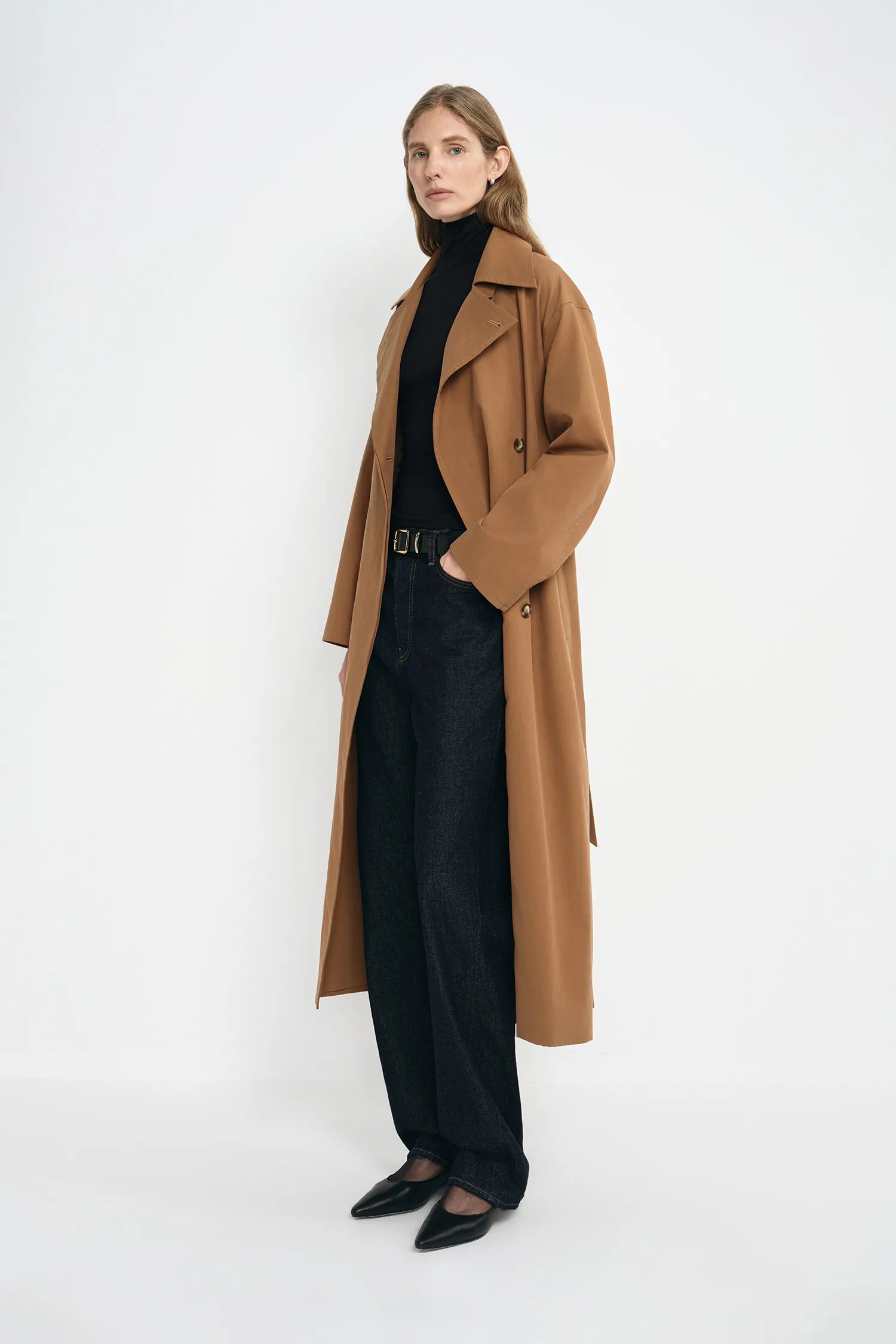 A model wearing a coppery brown oversized trench coat, pairing a black turtleneck and dark wash jeans, turned slightly to the side.