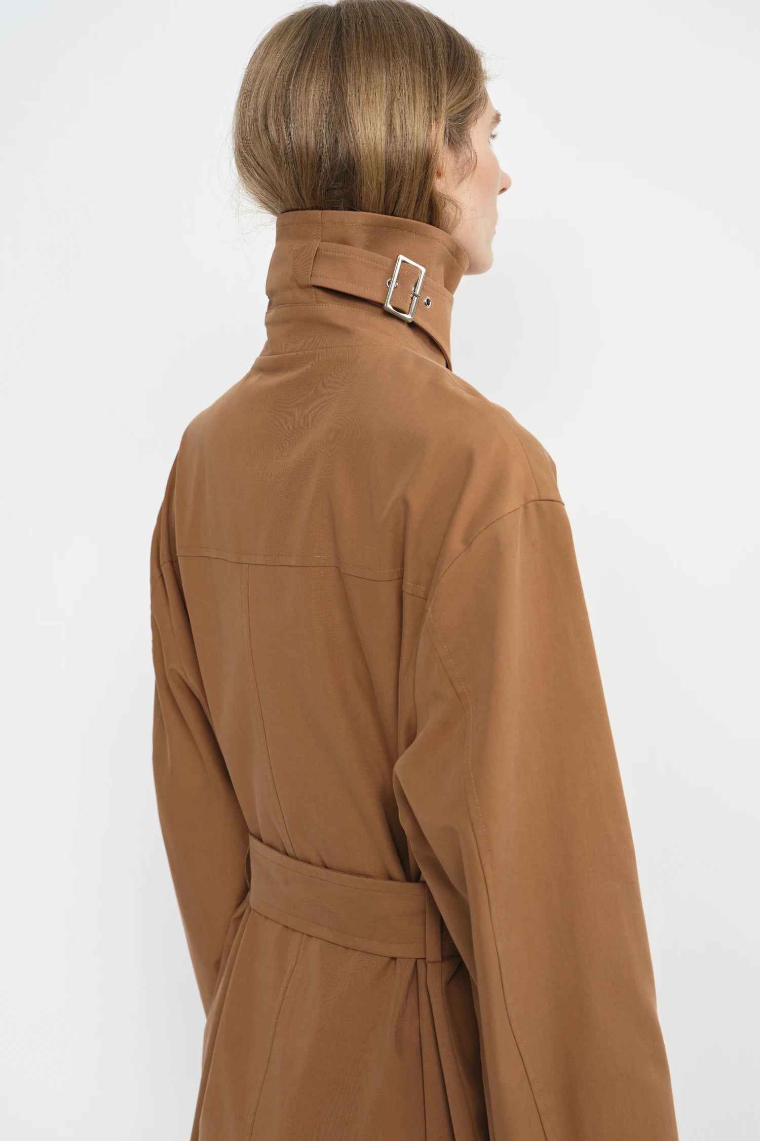 A model shown from the back wearing a camel brown trench coat with a high collar and a silver buckle detail, set against a plain white background.