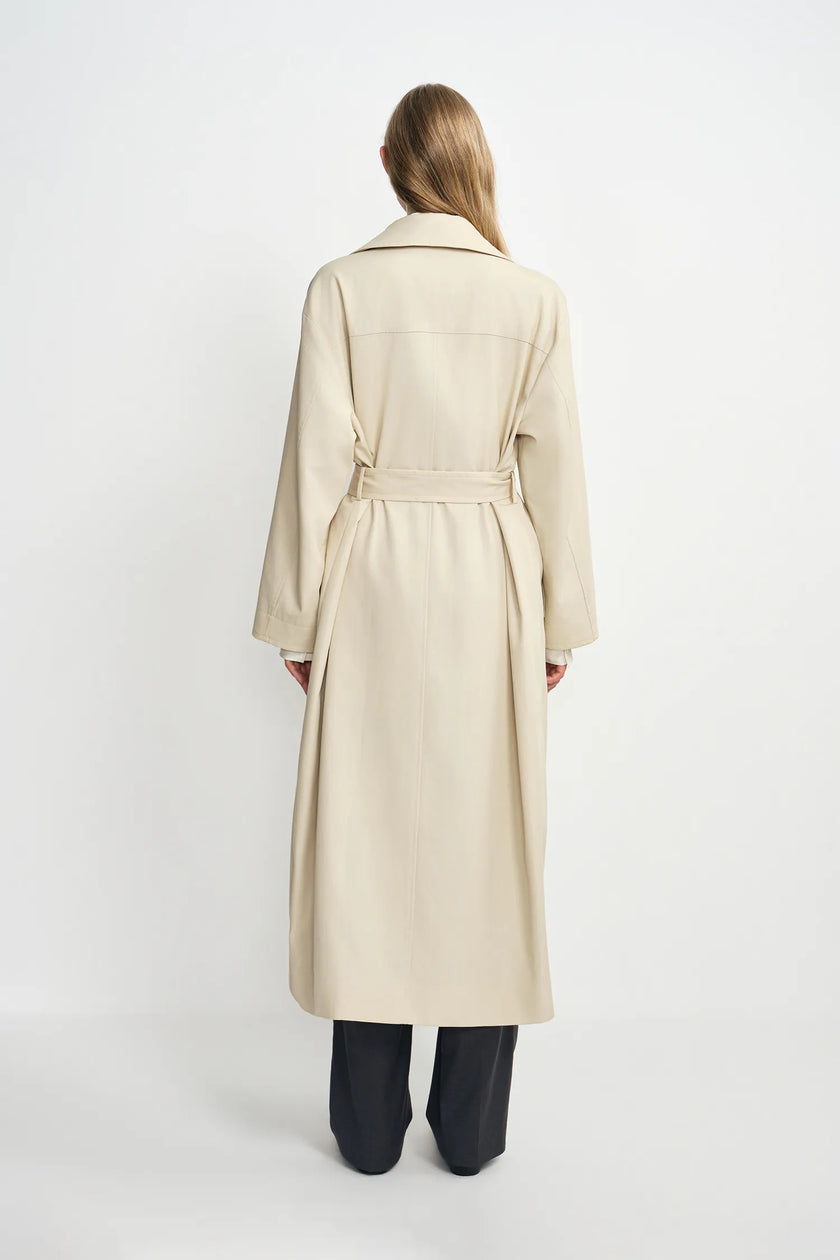 A model wearing an oatmeal oversized trench coat, pairing charcoal wide-leg trousers, back view.