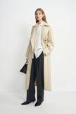 A model wearing an oatmeal oversized trench coat, open front, pairing charcoal wide-leg trousers, turned slightly to the side.
