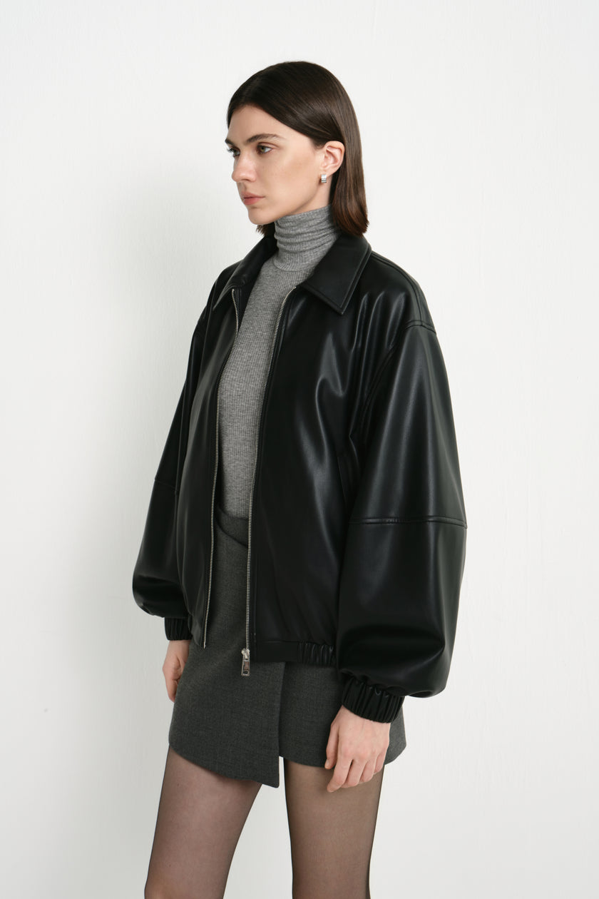 Side detail of a model in a black faux leather bomber, showing dropped shoulders and ballooning sleeves with gathered cuffs over a grey turtleneck and skirt.