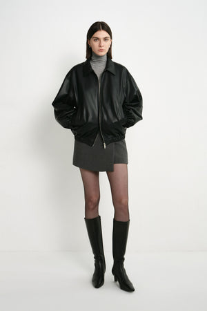 A model wearing a black faux leather bomber with oversized sleeves and a gathered hem, paired with a grey turtleneck, short grey skirt and black knee-high boots.
