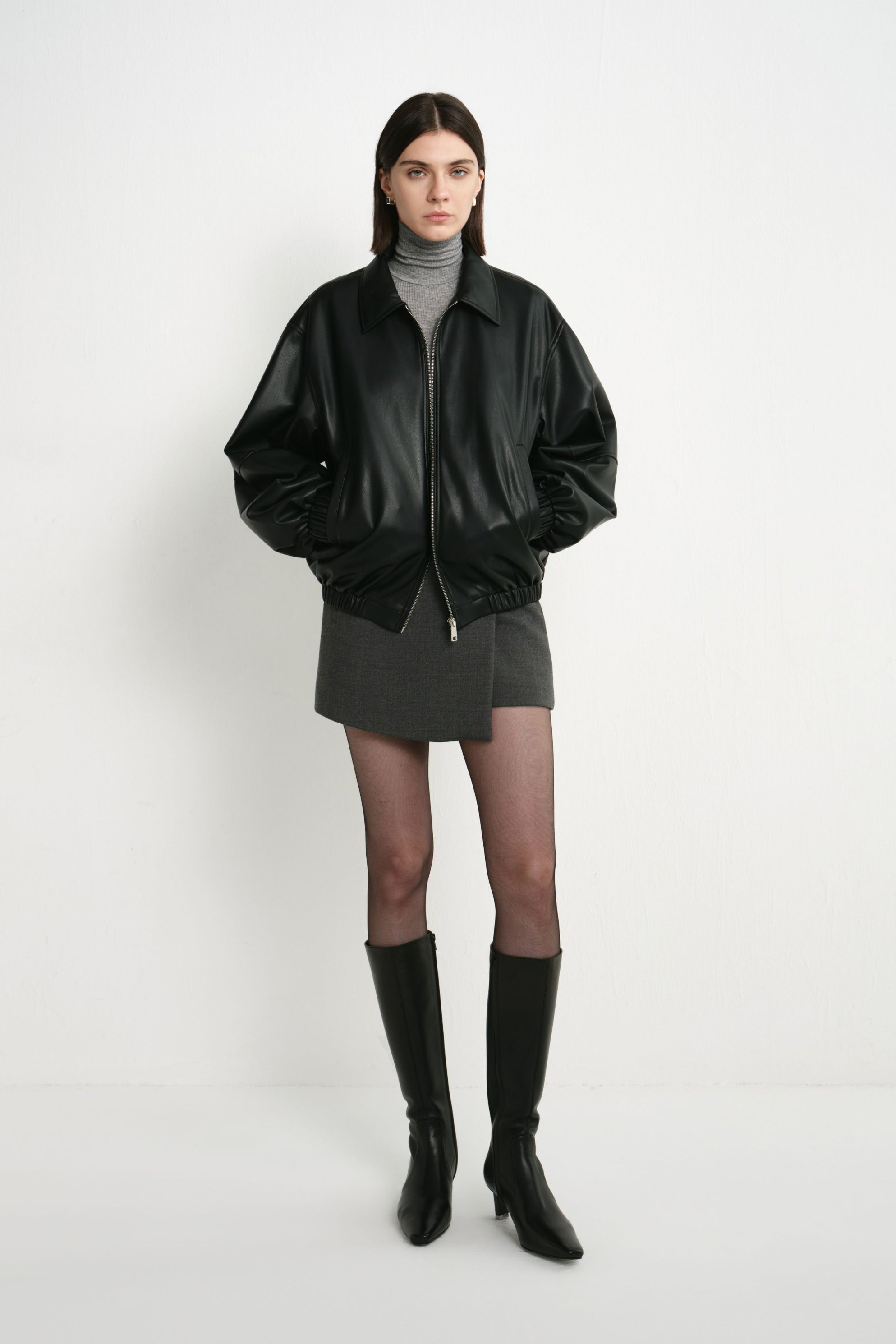 A model wearing a black faux leather bomber with oversized sleeves and a gathered hem, paired with a grey turtleneck, short grey skirt and black knee-high boots.