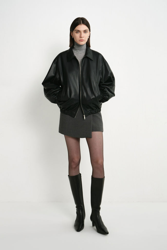 A model wearing a black faux leather bomber with oversized sleeves and a gathered hem, paired with a grey turtleneck, short grey skirt and black knee-high boots.
