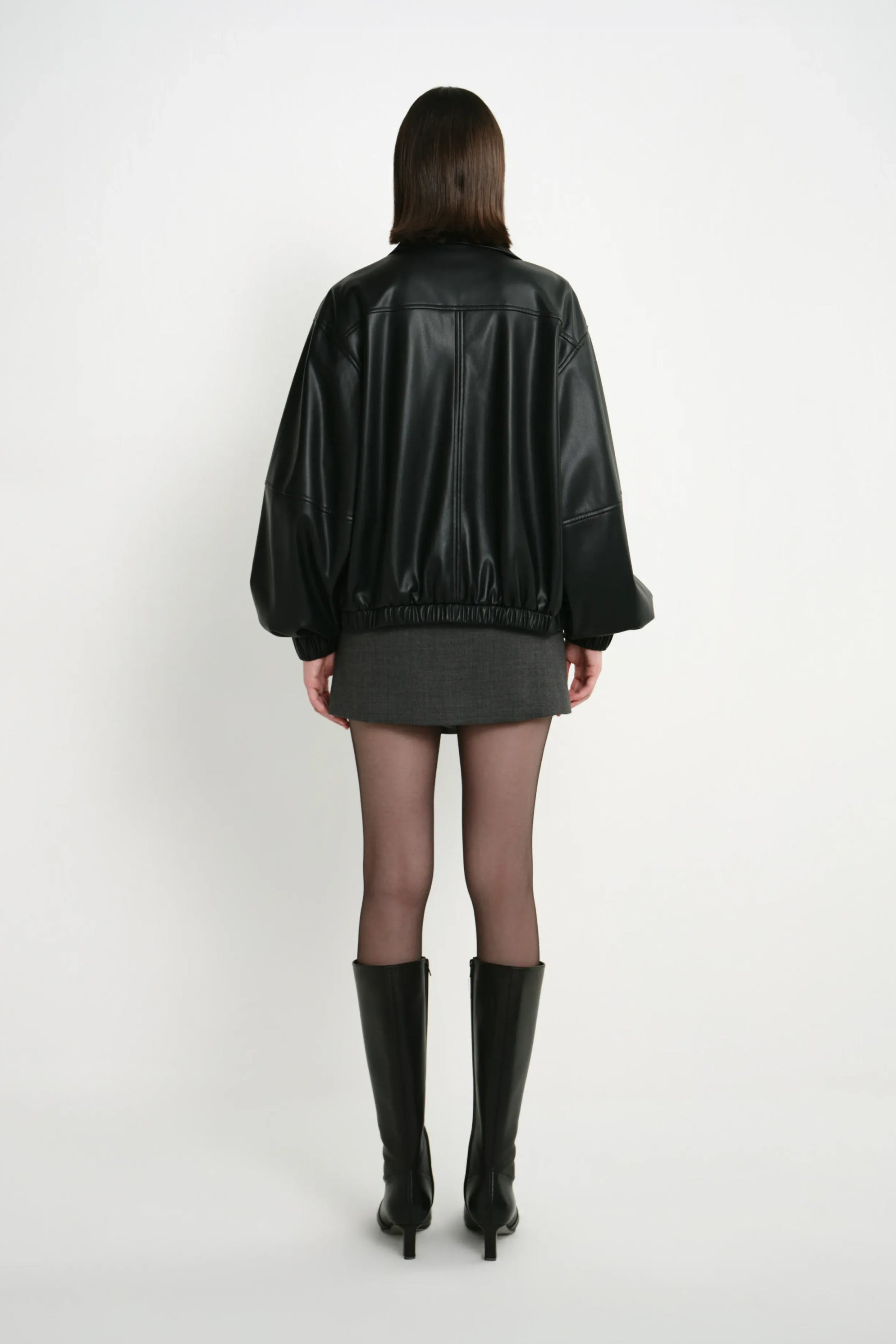 Back view of a model in a black faux leather bomber, showing the yoke seam, full sleeves and gathered hem above a grey skirt and black knee-high boots.