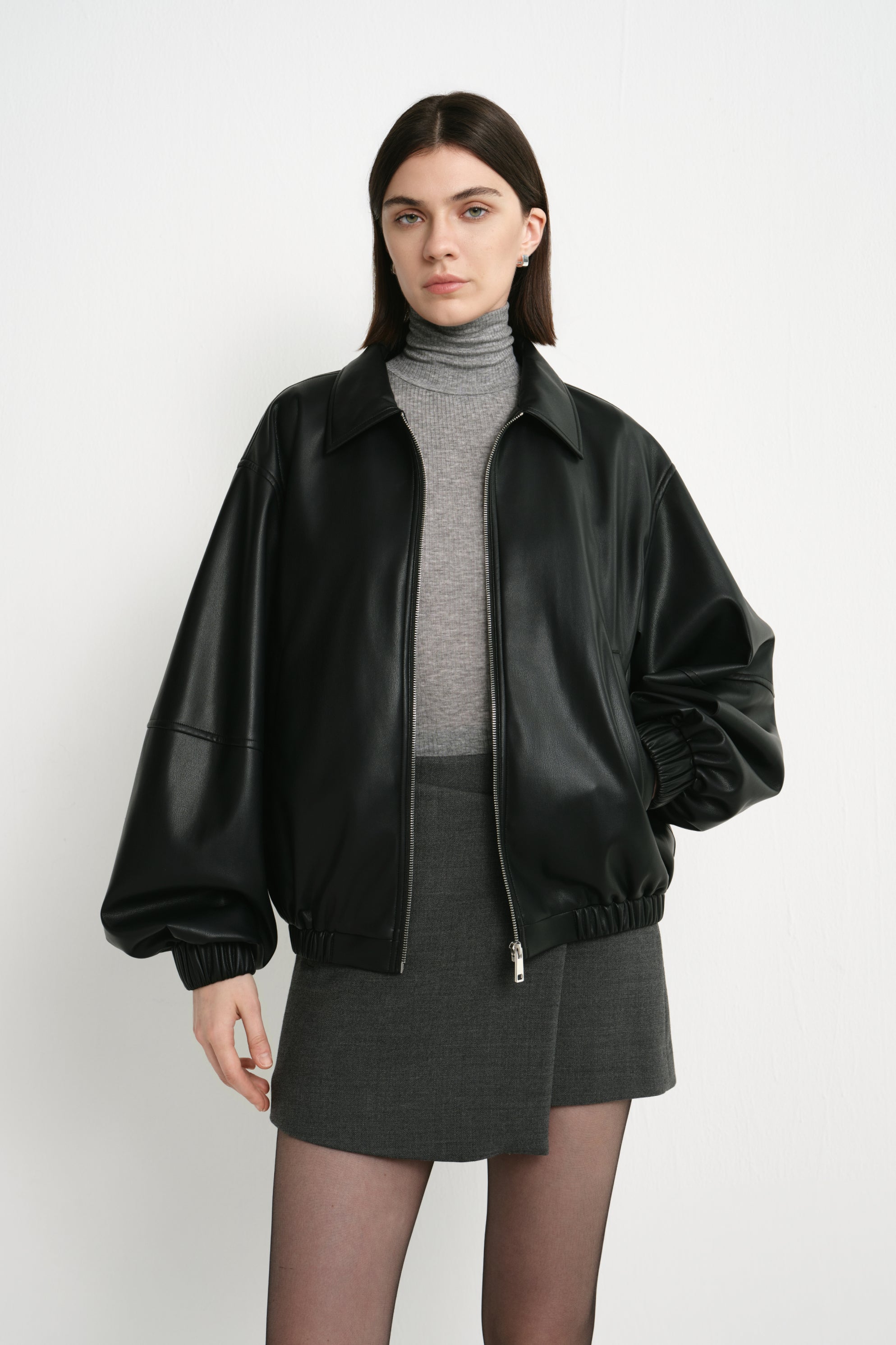 Front detail of a model wearing a black faux leather bomber, showing the pointed collar, broad sleeves and gathered hem over a grey turtleneck and skirt.