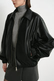 Close-up of a black faux leather bomber on a model, showing the pointed collar, metal zip and voluminous sleeve above a gathered cuff, over a grey turtleneck.