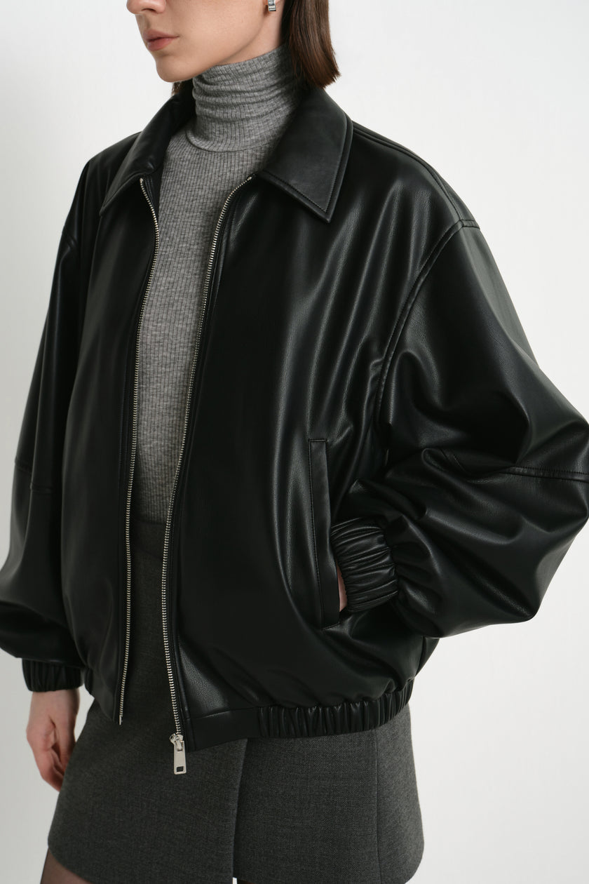Close-up of a black faux leather bomber on a model, showing the pointed collar, metal zip and voluminous sleeve above a gathered cuff, over a grey turtleneck.