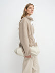 A woman in a beige knit zip-up sweater and cream trousers holds a large cream fabric clutch bag against a plain white studio background.