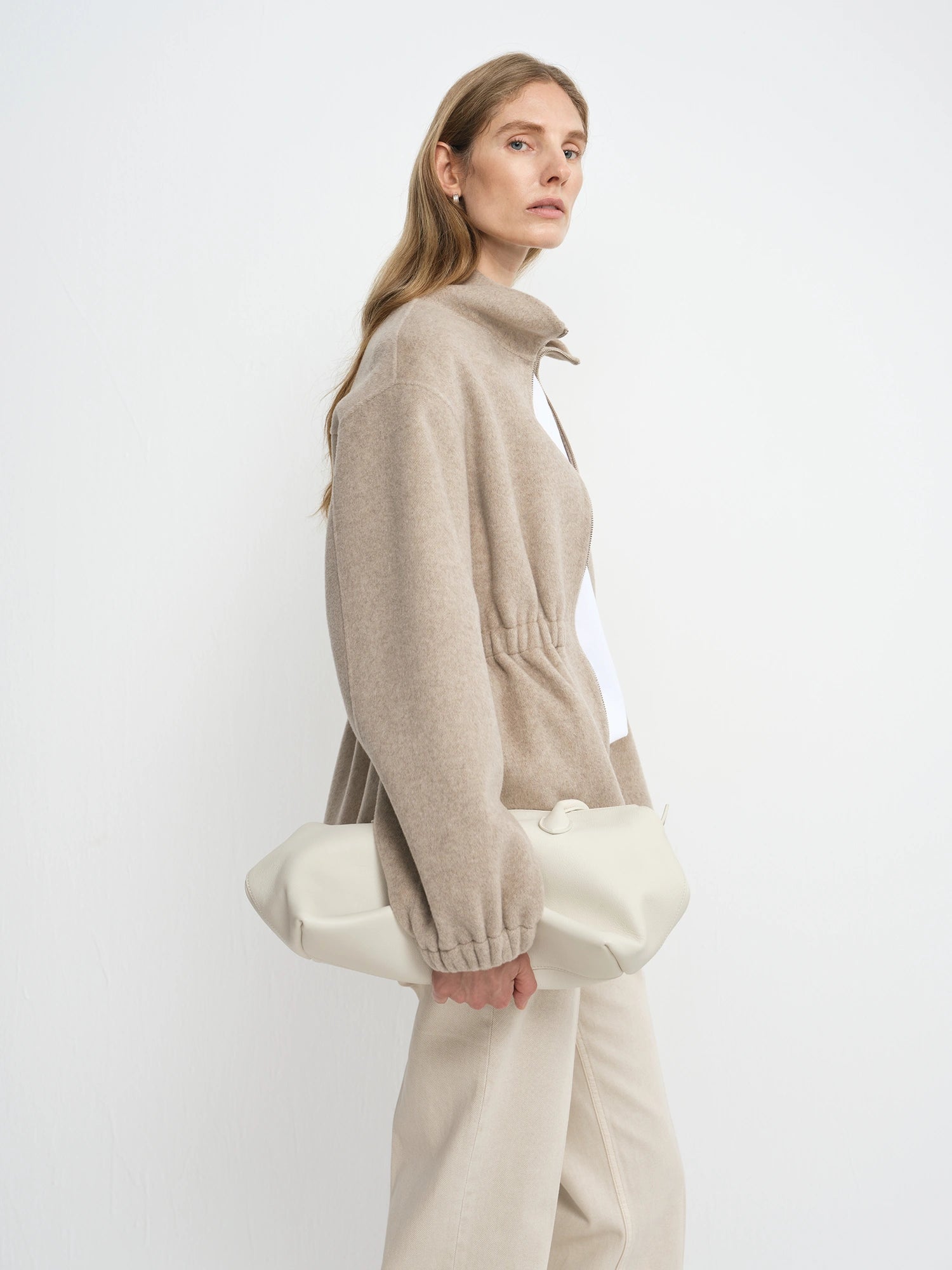 A woman in a beige knit zip-up sweater and cream trousers holds a large cream fabric clutch bag against a plain white studio background.