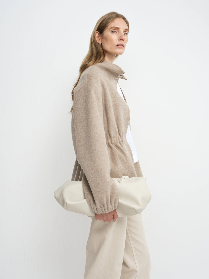 A woman in a beige knit zip-up sweater and cream trousers holds a large cream fabric clutch bag against a plain white studio background.