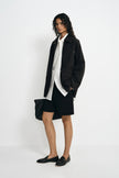 A model wearing a black utility jacket, pairing a white button-up shirt and black shorts, turned slightly to the side.