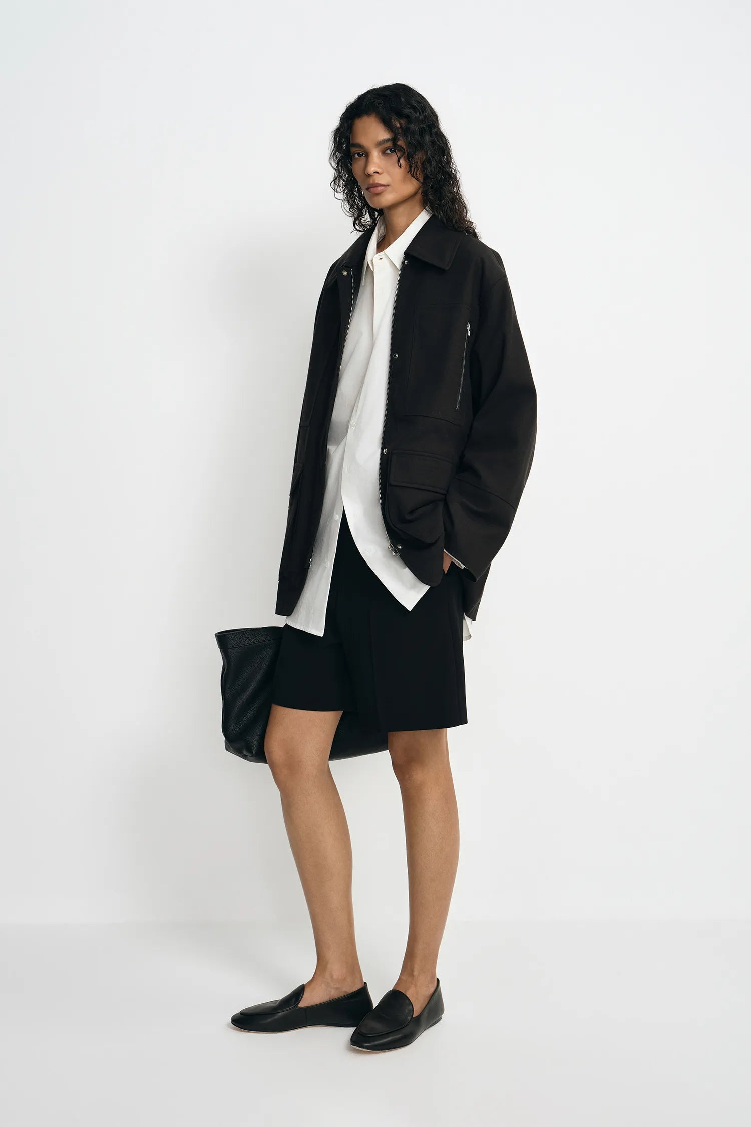 A model wearing a black utility jacket, pairing a white button-up shirt and black shorts, turned slightly to the side.