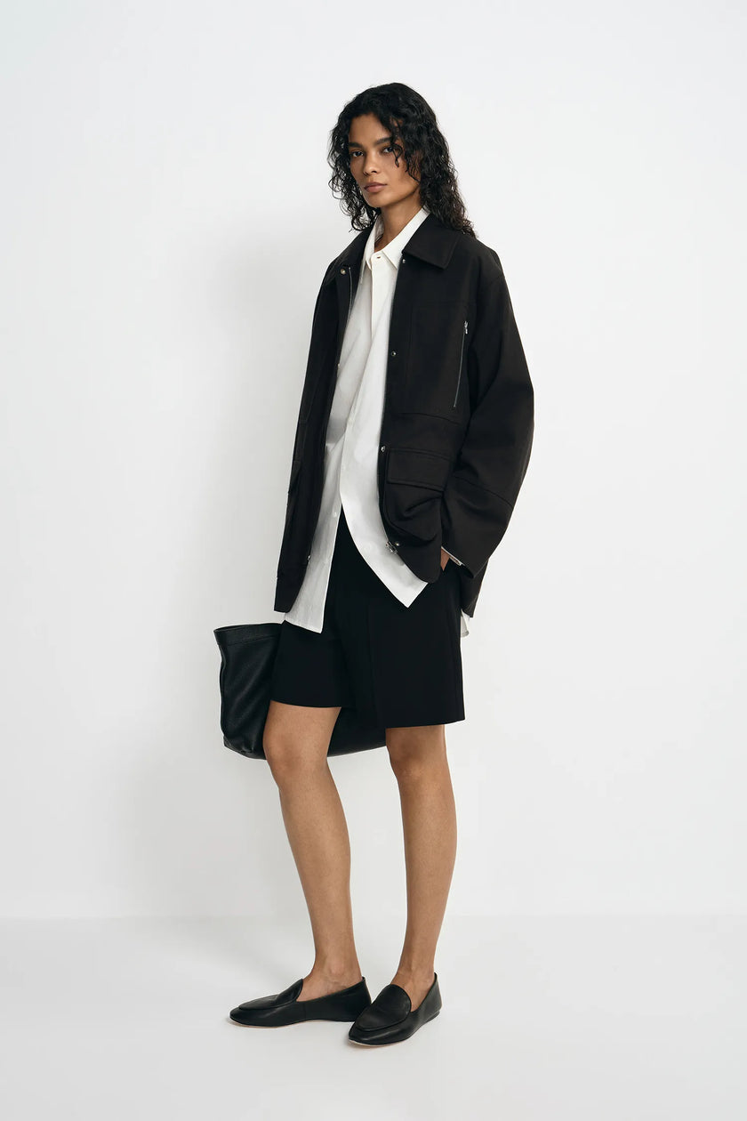 A model wearing a black utility jacket, pairing a white button-up shirt and black shorts, turned slightly to the side.