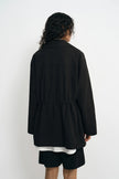 A model wearing a black utility jacket with a drawstring waist, back view.