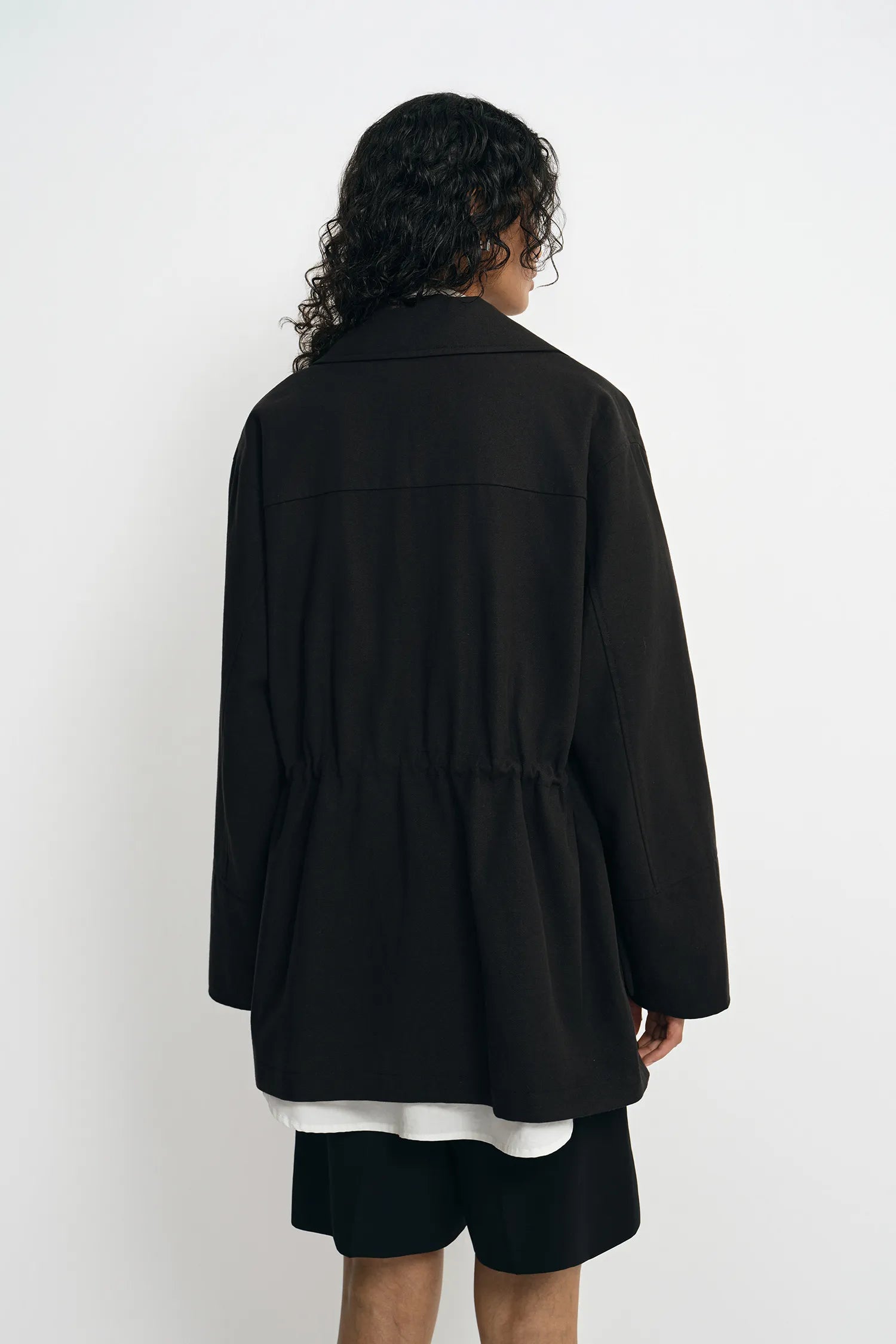 A model wearing a black utility jacket with a drawstring waist, back view.