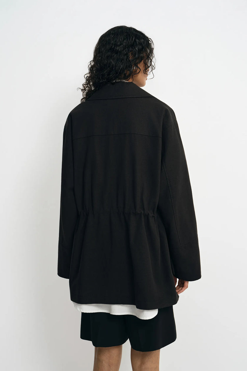 A model wearing a black utility jacket with a drawstring waist, back view.