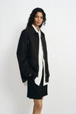A model wearing a black utility jacket, pairing a white button-up shirt and black shorts, side view.