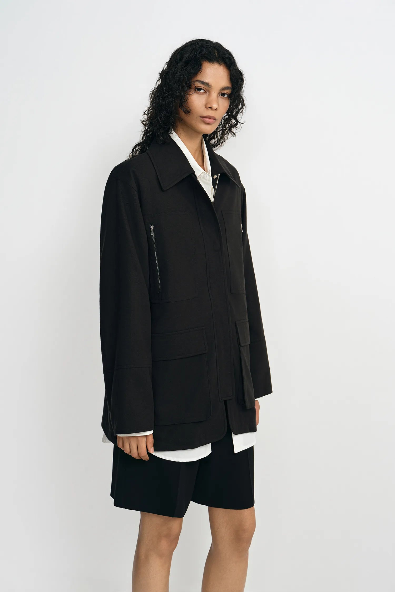 A model wearing a black utility jacket with zip pockets, turned slightly to the side.