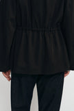 A close-up of a person wearing a black long-sleeve top with a gathered waist and dark pants, shown from the back against a plain light background.
