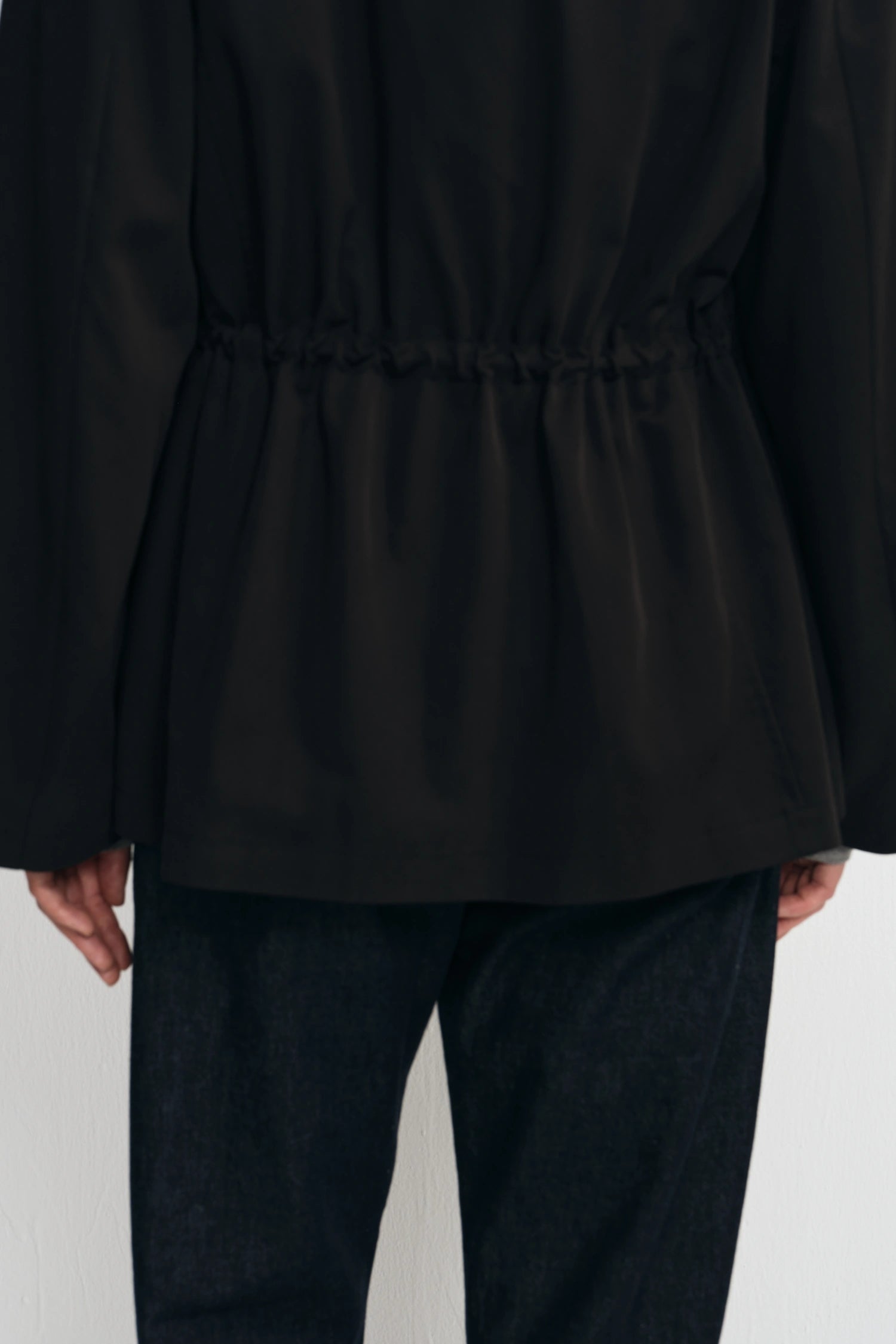 A close-up of a person wearing a black long-sleeve top with a gathered waist and dark pants, shown from the back against a plain light background.